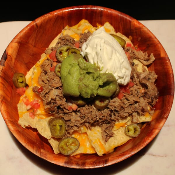 A wooden bowl of Kanaka nachos with shredded meat, cheese, jalapenos, guacamole, and sour cream.