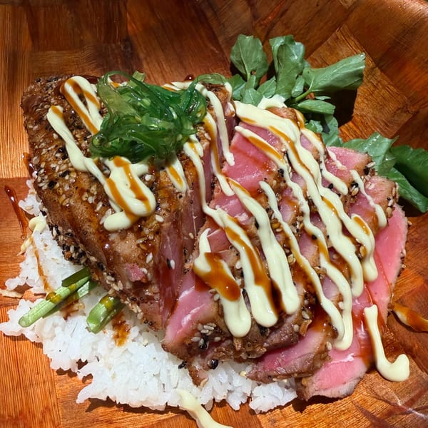 Seared tuna poke with sesame seeds, seaweed, and sauces, served over white rice in a wooden bowl.